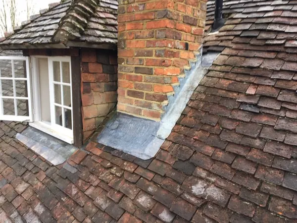 Lead flashing dressed down a valley where a brick chimney meets old peg tiles, beside a dormer window