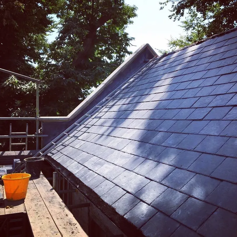 A finished slate roof on a steep pitch in sunlight, orange bucket and scaffold board at the eaves
